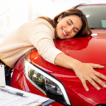 Consórcio Vale a Pena? Entenda Como Funciona Antes de Entrar 3 Uma mulher sorridente abraça o capô de um carro vermelho reluzente em uma concessionária. As chaves do carro, uma prancheta com documentos e um bloco de notas com uma marca de verificação verde estão sobre a mesa em primeiro plano.