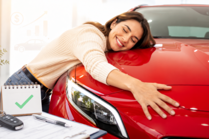 Uma mulher sorridente abraça o capô de um carro vermelho reluzente em uma concessionária. As chaves do carro, uma prancheta com documentos e um bloco de notas com uma marca de verificação verde estão sobre a mesa em primeiro plano.